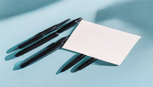 A flat-lay of three black ballpoint pens neatly arranged on a soft blue-grey paper background with a small folded white notecard, lit by quiet directional daylight.