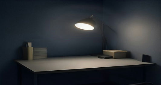 A desk lamp glowing over an empty desk in a dark blue room at dusk.
