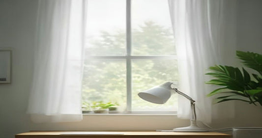 A bright home office corner with a white desk lamp on a wooden desk beside a window with sheer curtains and a leafy plant.