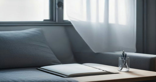 A grey sofa corner beside a small side table holding an open notebook and a glass of pens in soft window light.