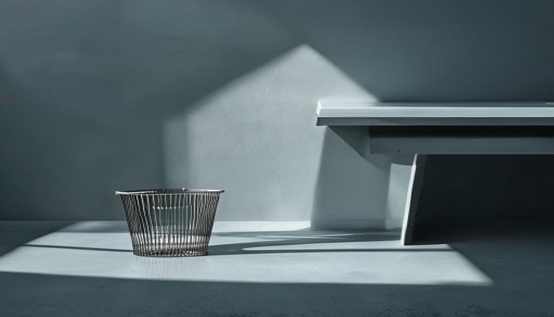 A slim brushed-metal mesh wastebasket beside a clean modern desk corner in cool slate-grey light.