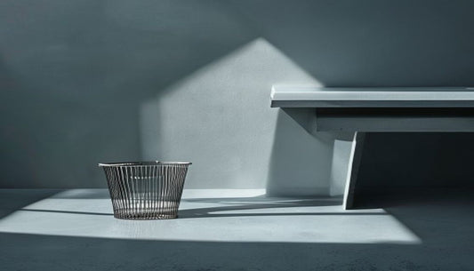 A slim brushed-metal mesh wastebasket beside a clean modern desk corner in cool slate-grey light.