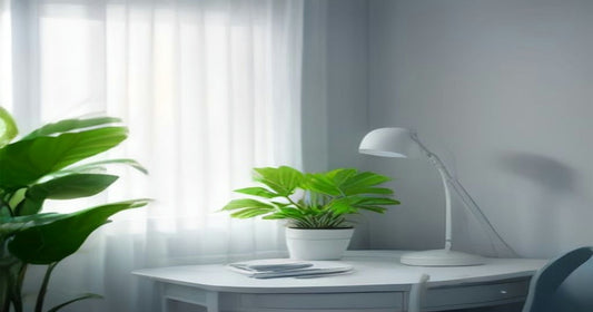 A white desk with a slim lamp and potted plant beside a sheer-curtained window in soft cool daylight.