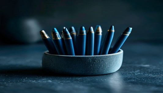 A small cluster of pens standing in a matte ceramic holder on a dark slate desk, lit dramatically from the side in cool blue-grey tones.